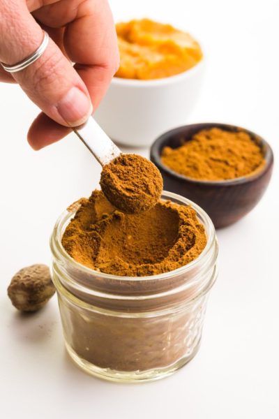 A hand holds a measuring spoon full of pumpkin pie spice over a bowlful of the spice. There's a bowl of pumpkin puree and another bowl of spice in the background.