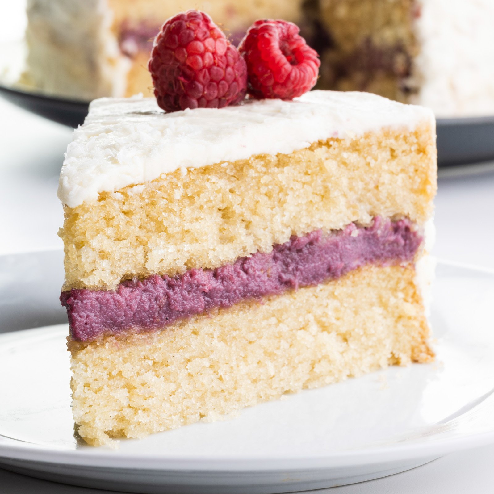 A slice of raspberry coconut cake on a plate sits in front of the rest of the cake.