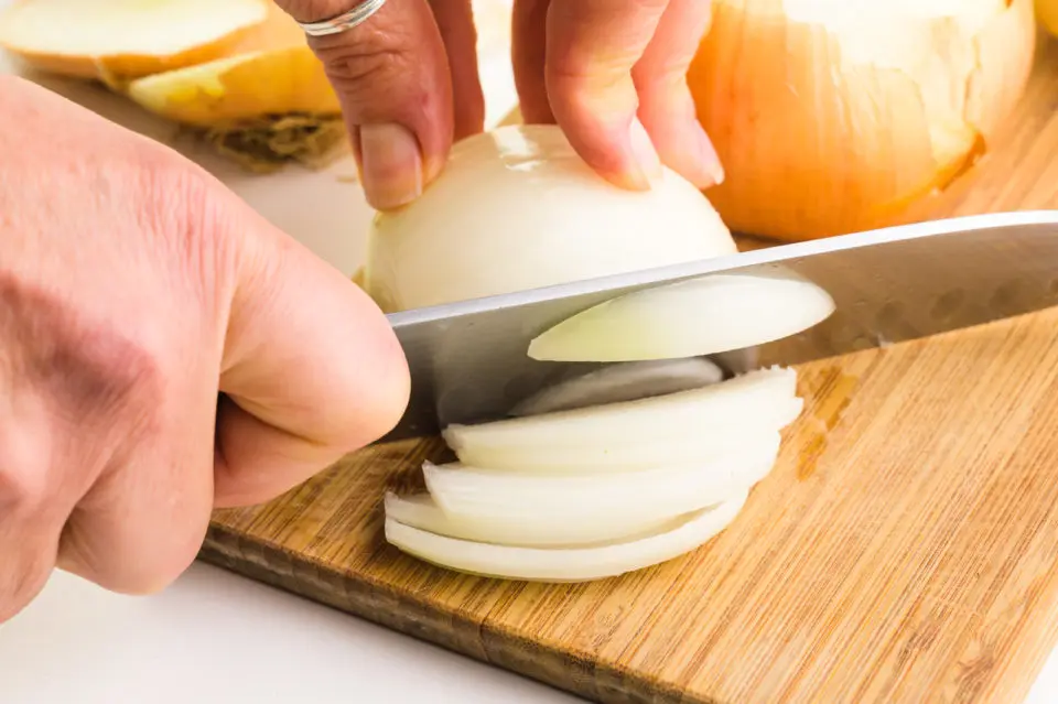 A hand holds a knife and is slicing an onion on cutting board. There are other onions around it.