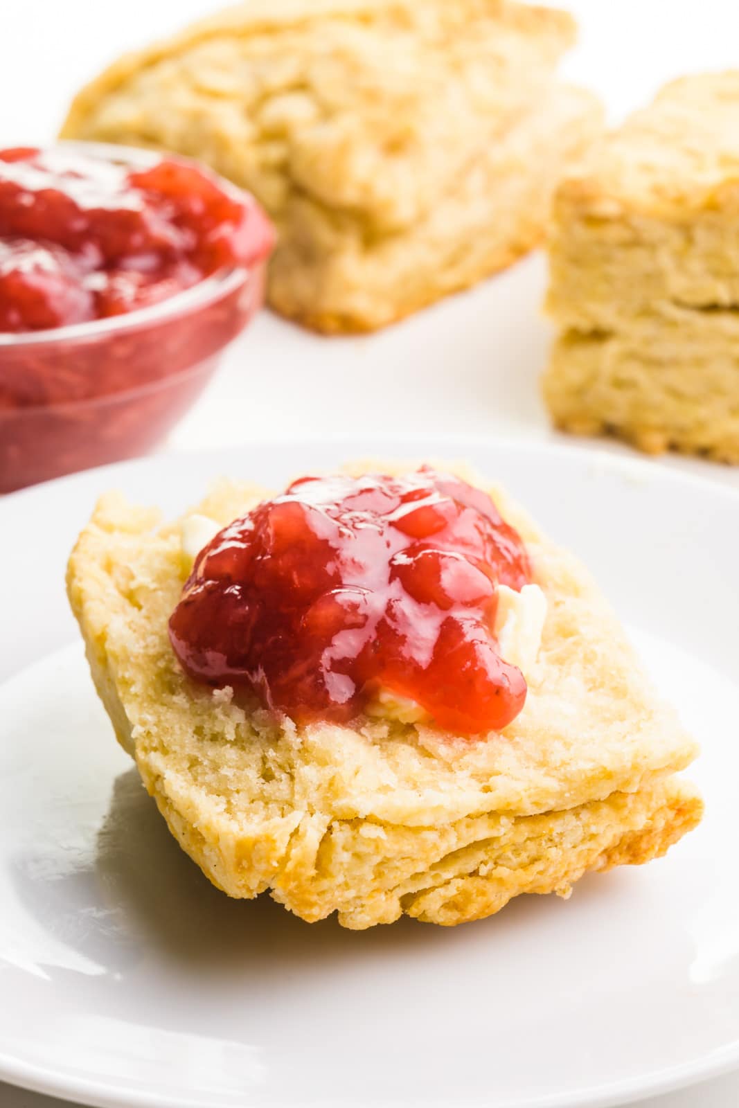 Half of a biscuit is on a plate. It's topped with butter and strawberry jelly. There's a bowl with more jelly and more biscuits behind it.