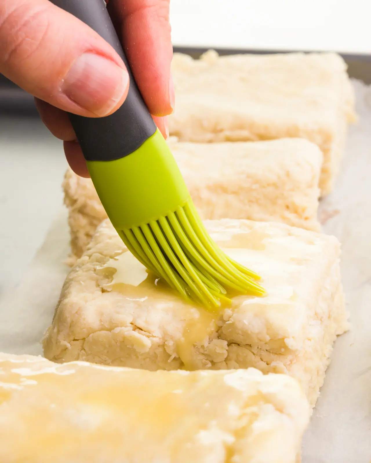 A hand holds a pastry brush and is spreading vegan egg wash over biscuit dough.
