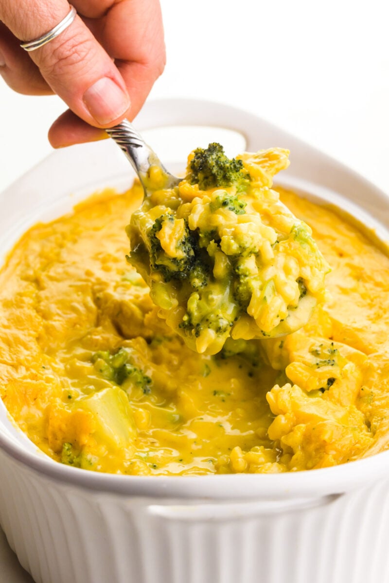 A hand holds a spoonful of vegan broccoli cheese casserole over the rest of the casserole dish.