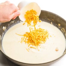Cheese is being poured into a skillet with a creamy mixture in it.