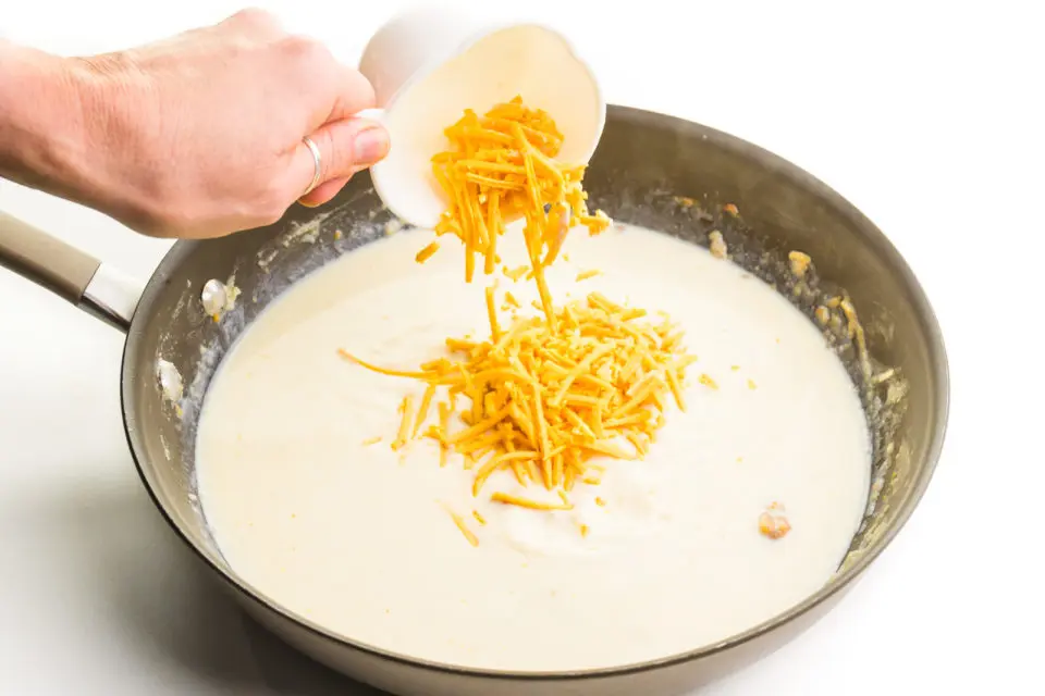 Cheese is being poured into a skillet with a creamy mixture in it.