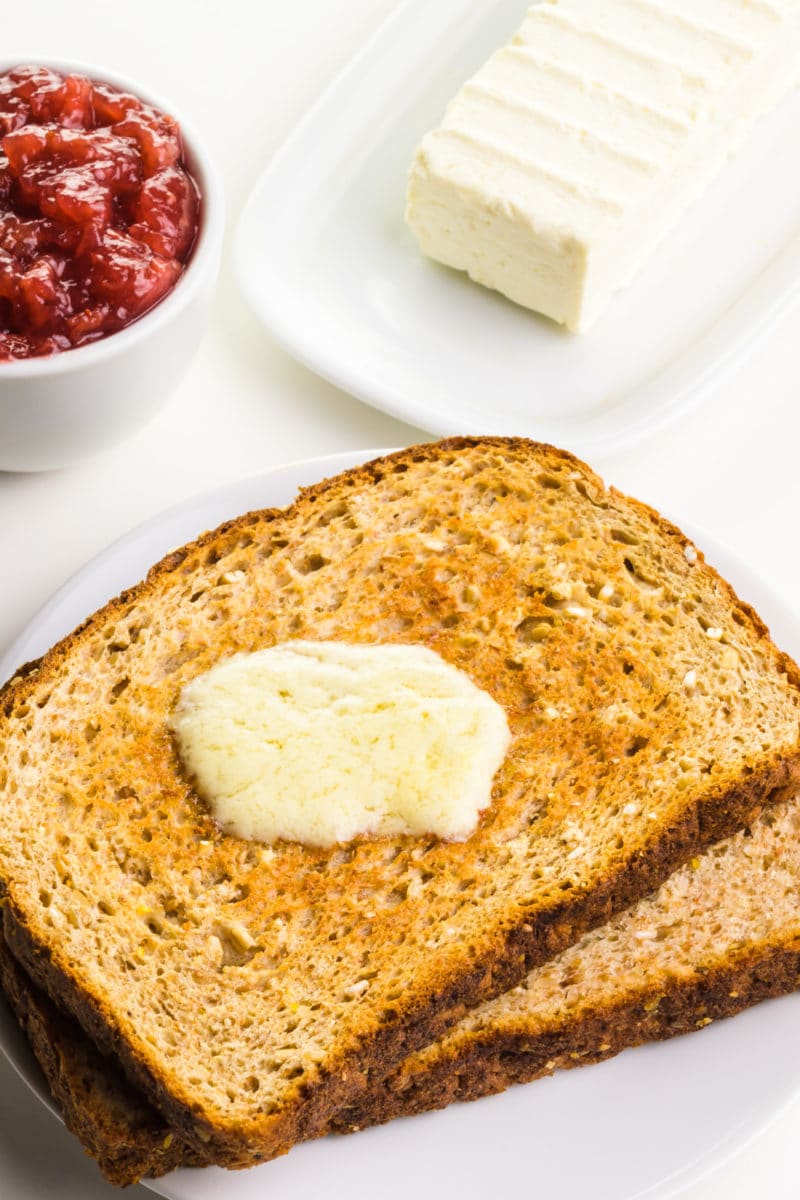 Slices of toast are stacked on a plate. The top piece has melted vegan butter on top. There's a bowl of jelly and more butter on a plate behind it.