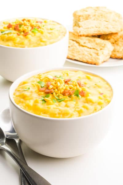 A bowl of corn chowder has spoons next to it. There's another bowl of chowder and biscuits in the background.