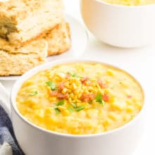 Two bowls of vegan corn chowder have vegan bacon and cheese on top. There's a blue towel next to the front bowl and a stack of biscuits in the background.