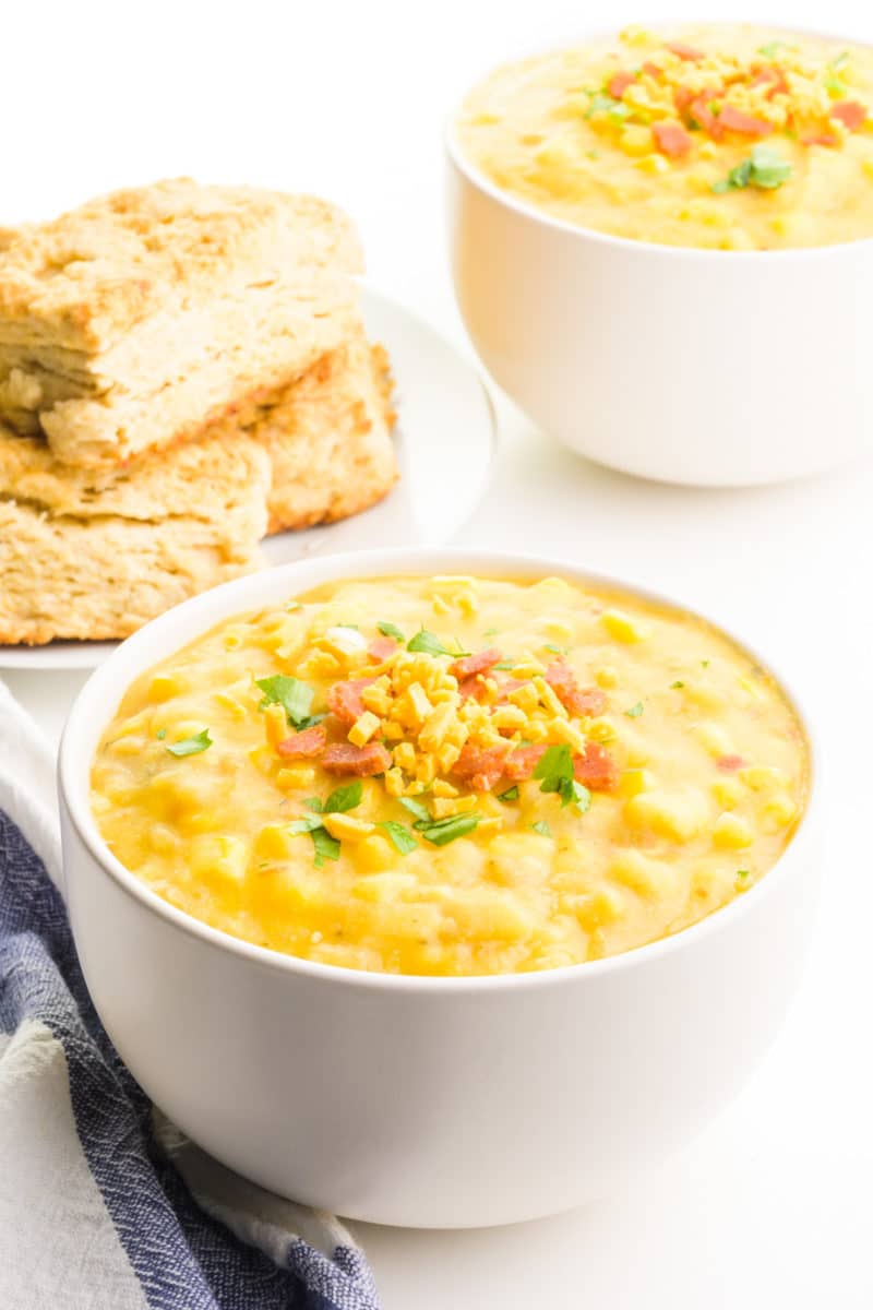 Two bowls of vegan corn chowder have vegan bacon and cheese on top. There's a blue towel next to the front bowl and a stack of biscuits in the background.