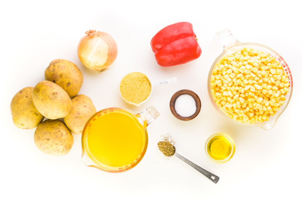 Ingredients sit on a white background, including golden potatoes, an onion, a red bell pepper, a bowl of frozen corn, broth, and more.