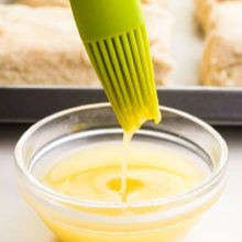A silicone pastry brush is dipped in a bowl with vegan egg wash. A pan of pastries sits behind it.
