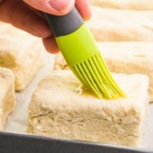 A hand holds a pastry brush, brushing liquid over unbaked biscuits.