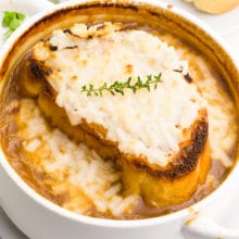 A bowl of vegan French onion soup has melted cheese on top. There's sprigs of fresh herbs behind it and toasted baguette slices.