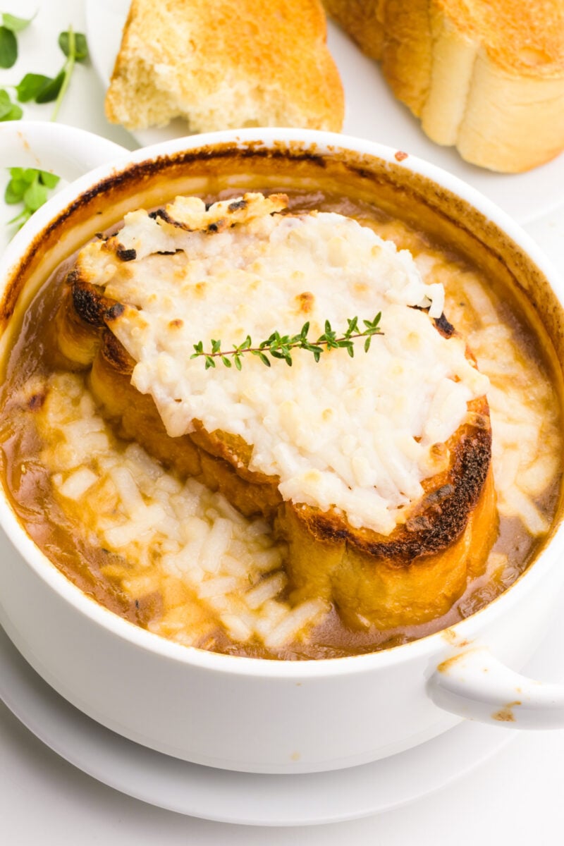 A bowl of vegan French onion soup has melted cheese on top. There's sprigs of fresh herbs behind it and toasted baguette slices.