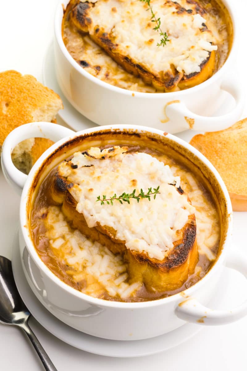 Two bowls of onion soup has melted cheese on top. There are slices of toasted baguette around them and a spoon by the front bowl.