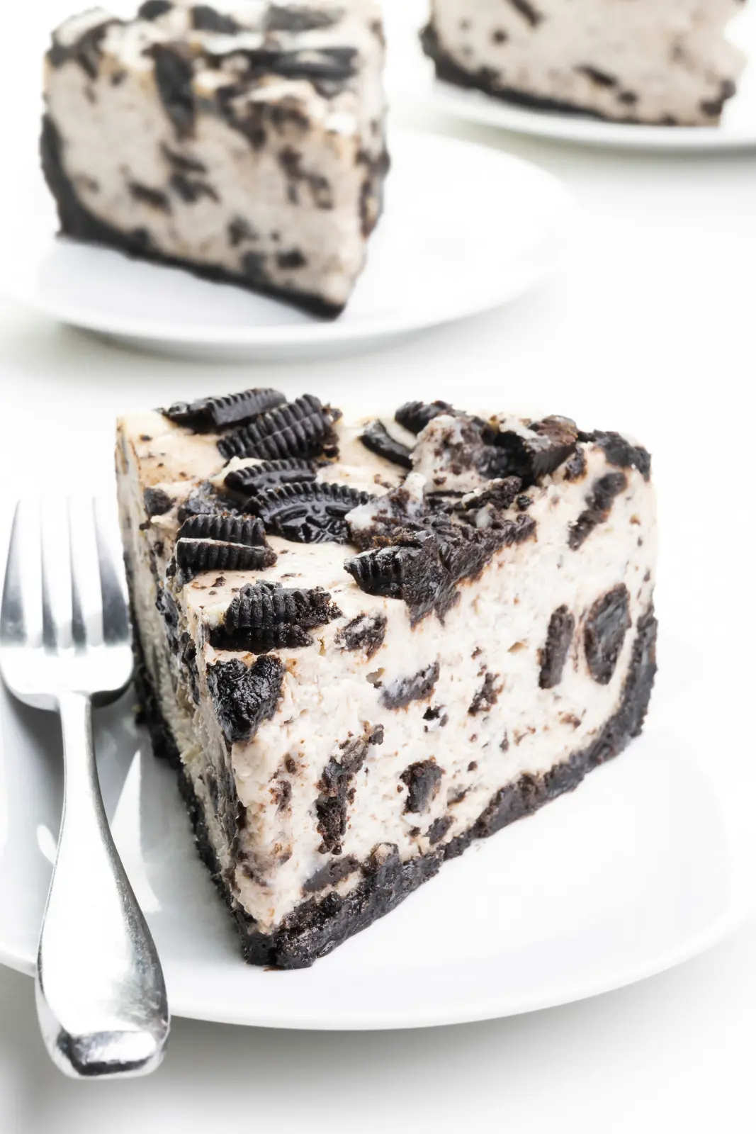 A slice of Oreo cheesecake sits on a plate with a fork beside it. There are two more slices on plates behind it.