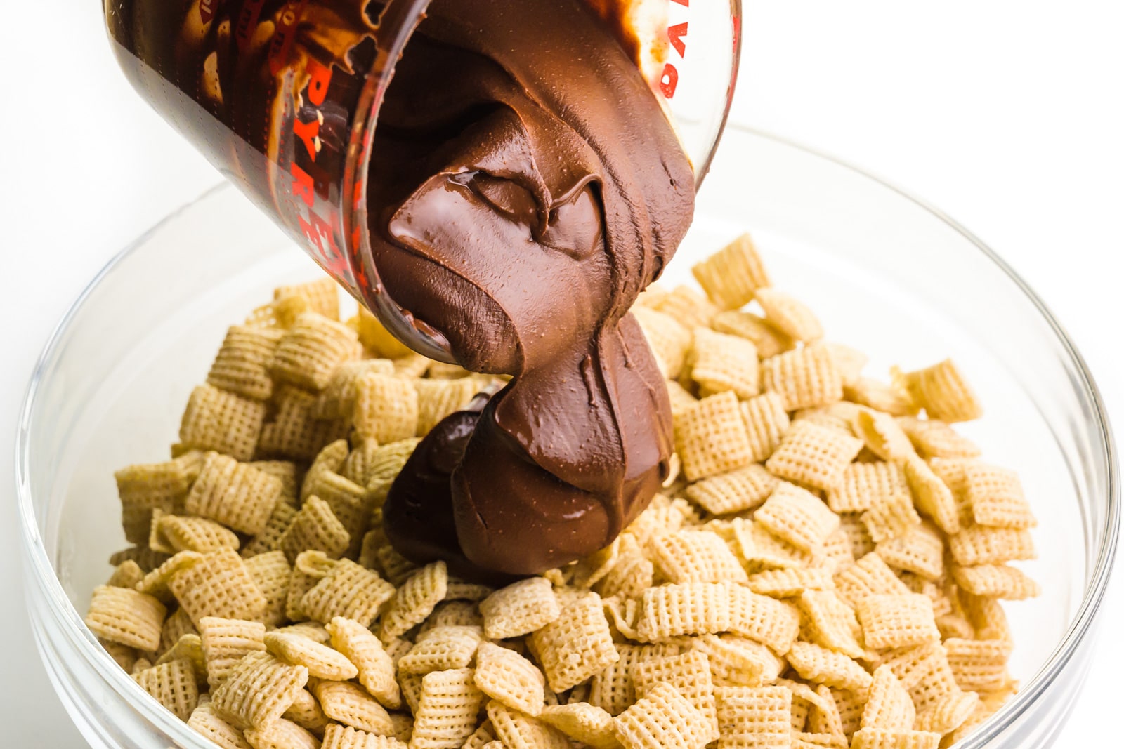 A chocolate mixture is being poured into a bowl full of Chex cereal.