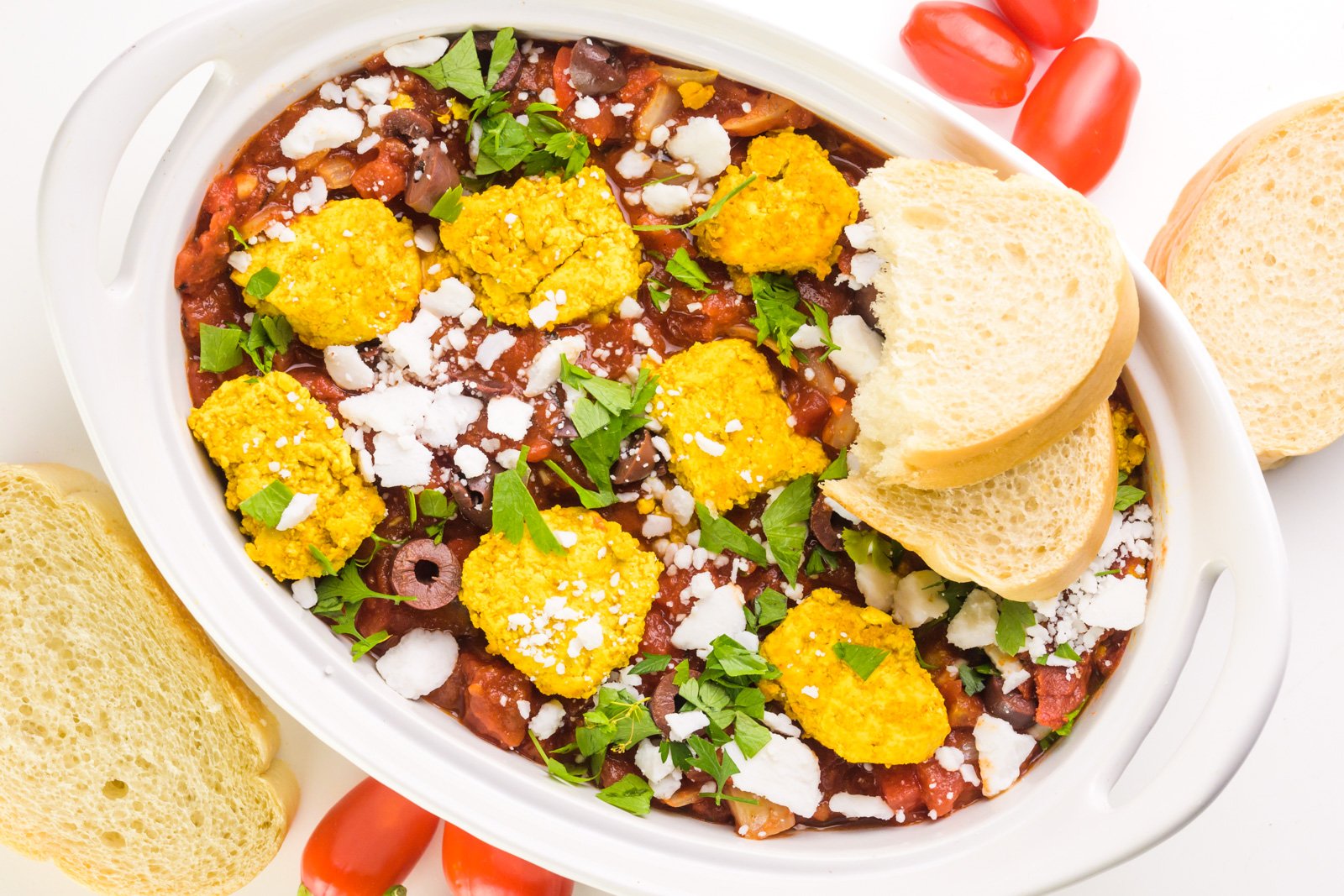 Looking down on a Mediterranean casserole with tofu and vegan feta crumbles. There are two slices of bread on the dish and mini red bell peppers beside it.