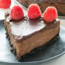 A slice of vegan chocolate cheesecake has a row of fresh raspberries on top. The rest of the cheesecake is sitting behind the slice.