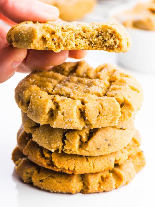 A stack of vegan peanut butter cookies is sitting in the foreground. A hand is holding a cookie right above the stack and there's a bite taken out of that cookie.