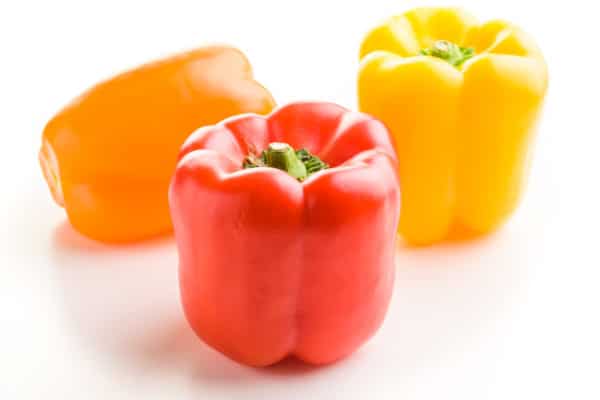 Three bell peppers sit on a white counter. One is red, the other is orange, the the other one is yellow.