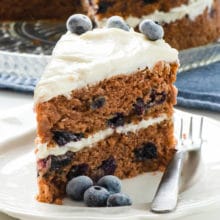 A slice of 2-layer vegan blueberry cake with fresh blueberries on top sits in front of the whole cake.