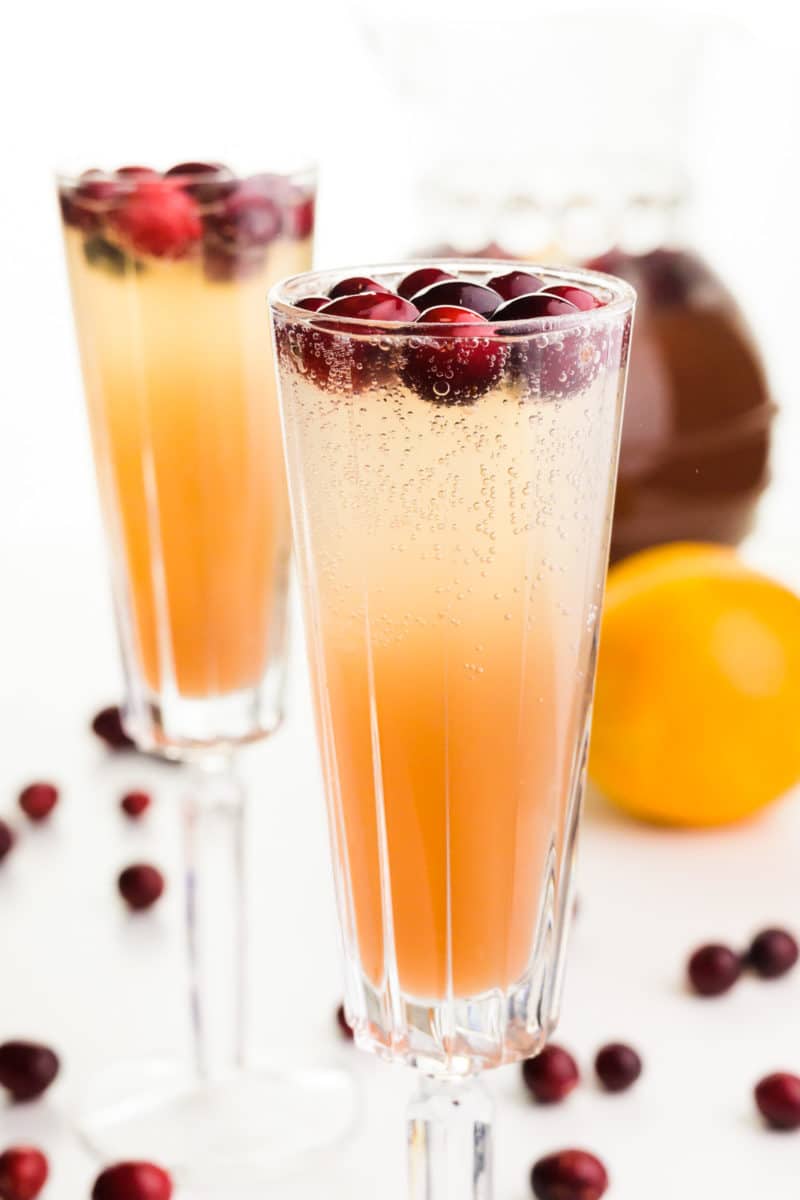 Two long-stemmed glasses are full of Christmas punch with fresh cranberries. The pitcher of punch concentrate is behind it along with a fresh orange and fresh cranberries.
