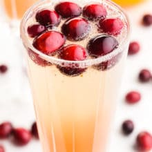 A close-up image of a glass of holiday punch shows fresh cranberries and sparkling water fizzes at the tope. There are fresh cranberries, an orange, and another glass in the background.