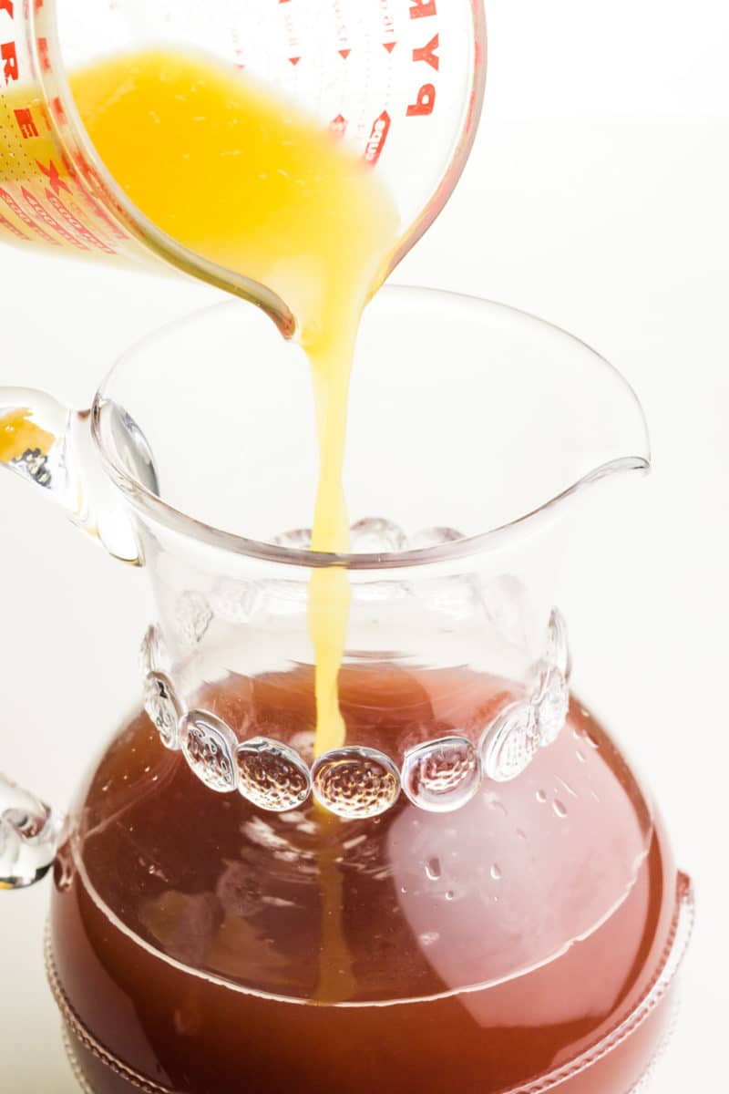 Orange juice is being poured into a pitcher with other fruit juices.