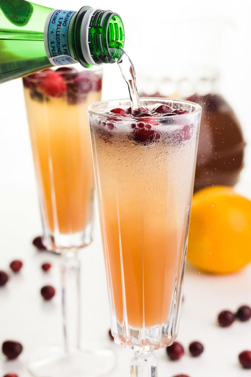 Sparkling water is being poured into a glass full of punch and fresh cranberries. There's another glass, a pitcher, and fresh cranberries behind it.