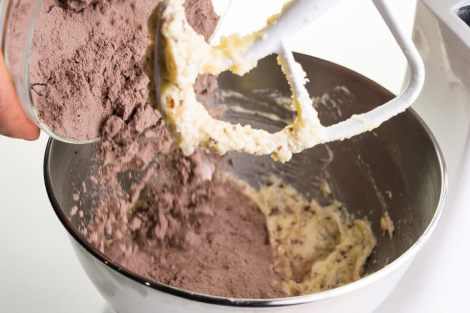 A chocolate flour mixture is poured into a stand mixer bowl with a creamed butter mixture.