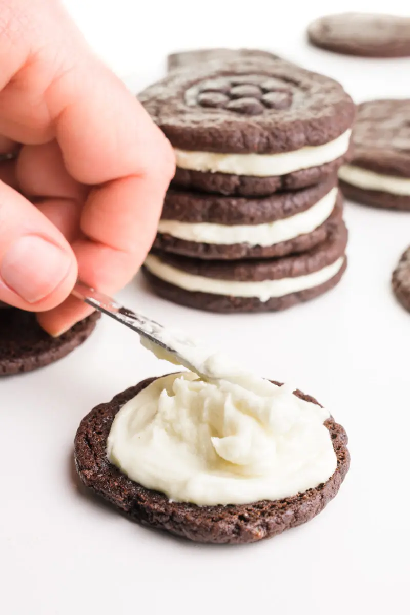A hand is spreading homemade Oreo filling over a chocolate cookie to make homemade Oreos. There are more of the chocolate sandwich cookies in the background.