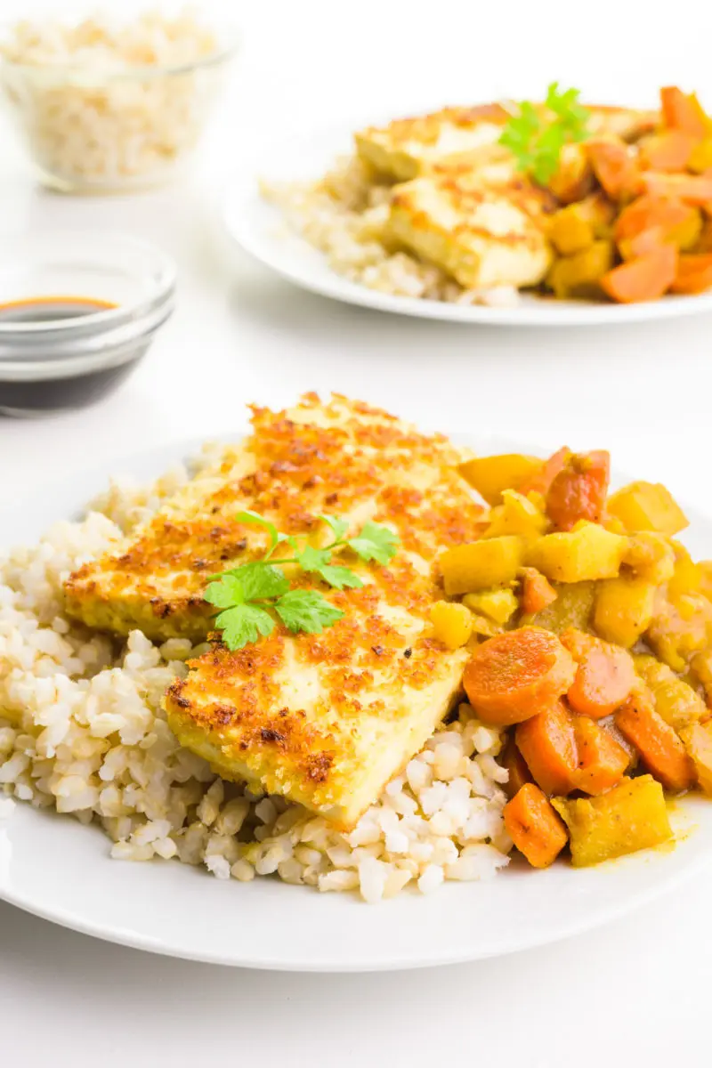 Fried tofu sits on a bed of rice with a vegetable sauce beside it. There are bowls of soy sauce and rice behind it and another plate.