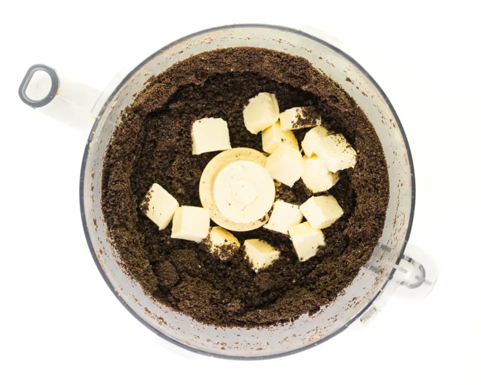 Cookie crumbs are in a food processor bowl with cubes of vegan butter on top.