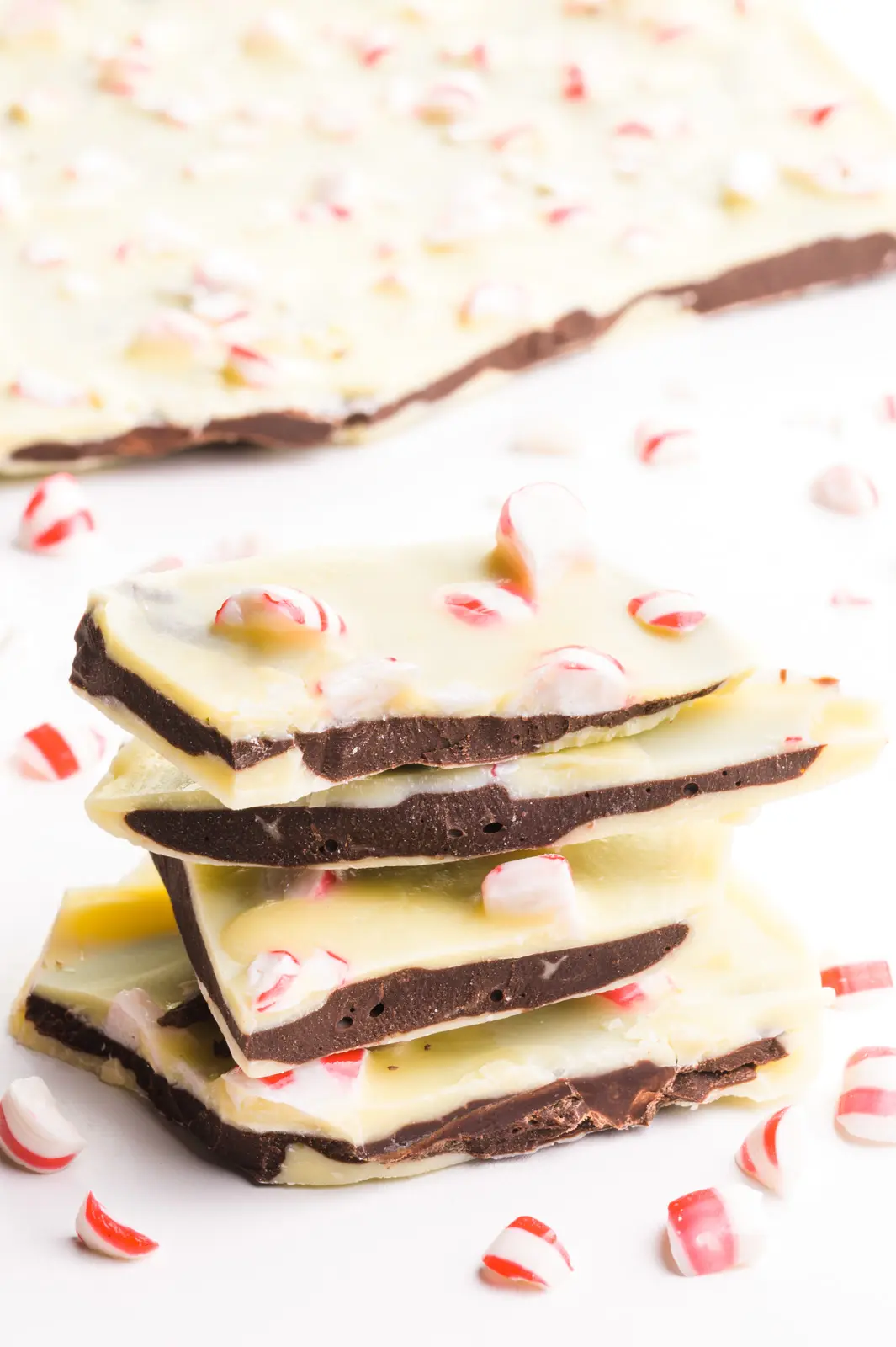 A stack of vegan peppermint bark has peppermint pieces around it and a block of bark behind it.