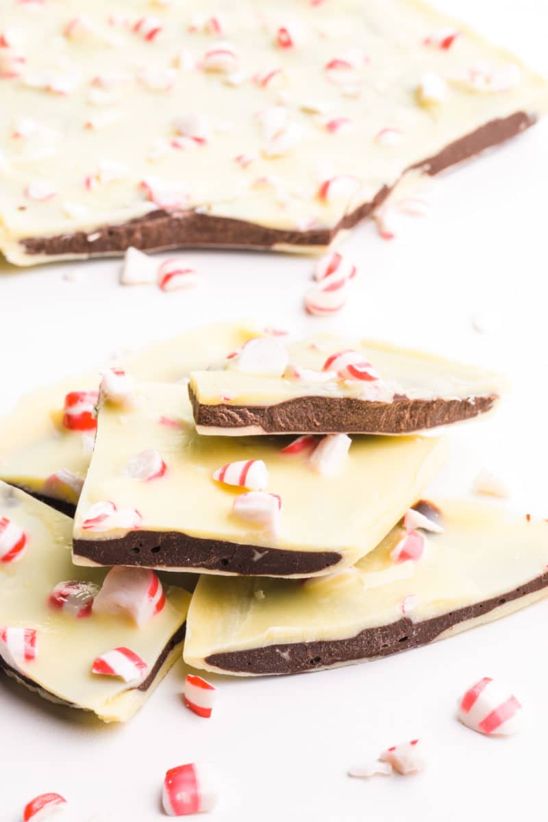A stack of peppermint bark has peppermint candy pieces around it. It sits in front of a big block of bark.