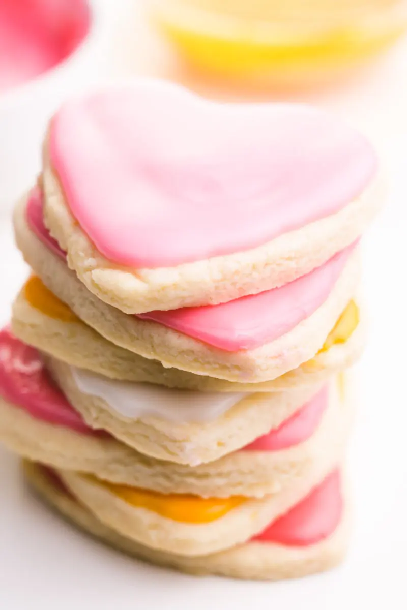 A stack of heart-shaped sugar cookies have pink and orange frosting. There are bowls of frosting behind them.