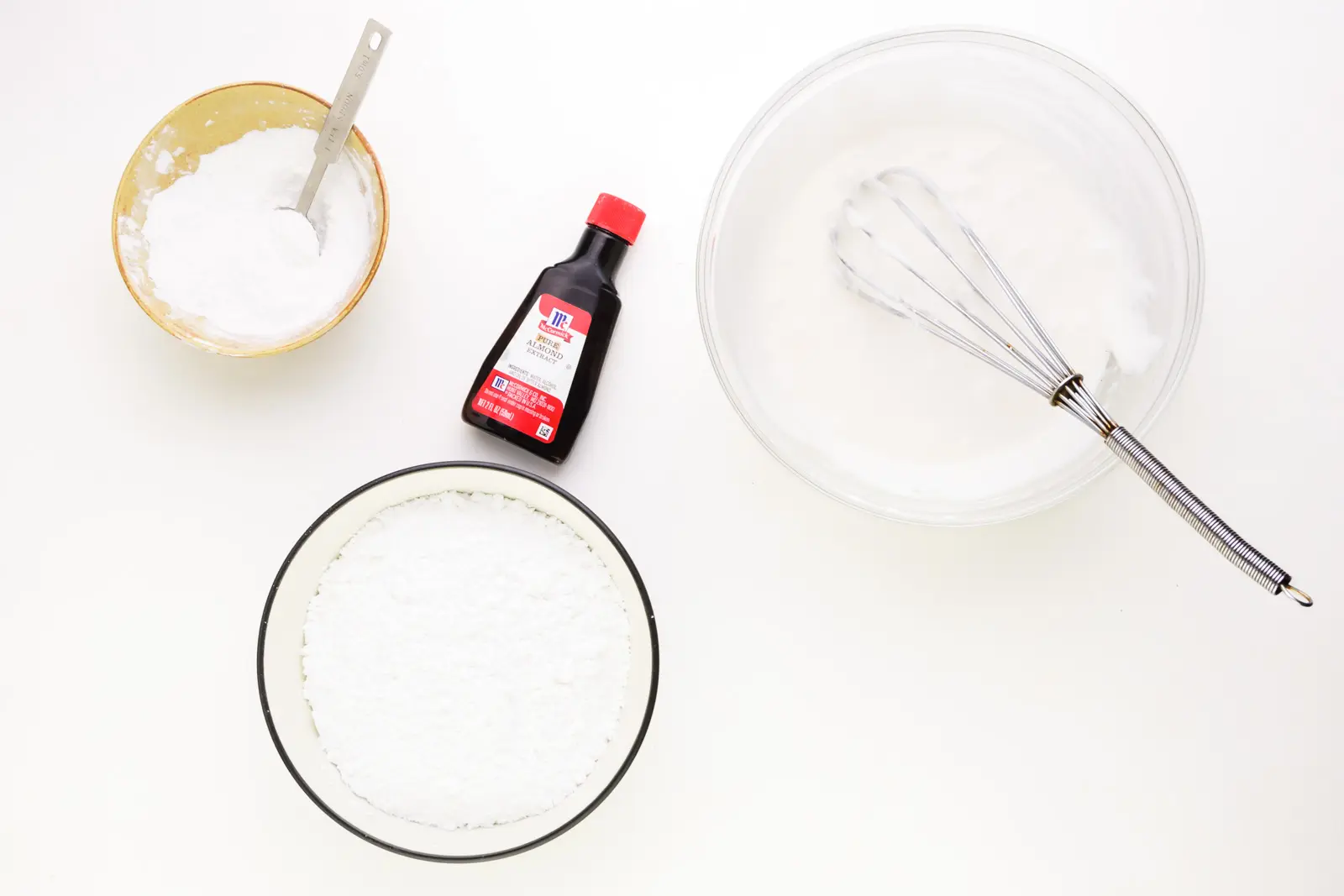Ingredients for frosting sit on a white counter, including powdered sugar, and almond extract.