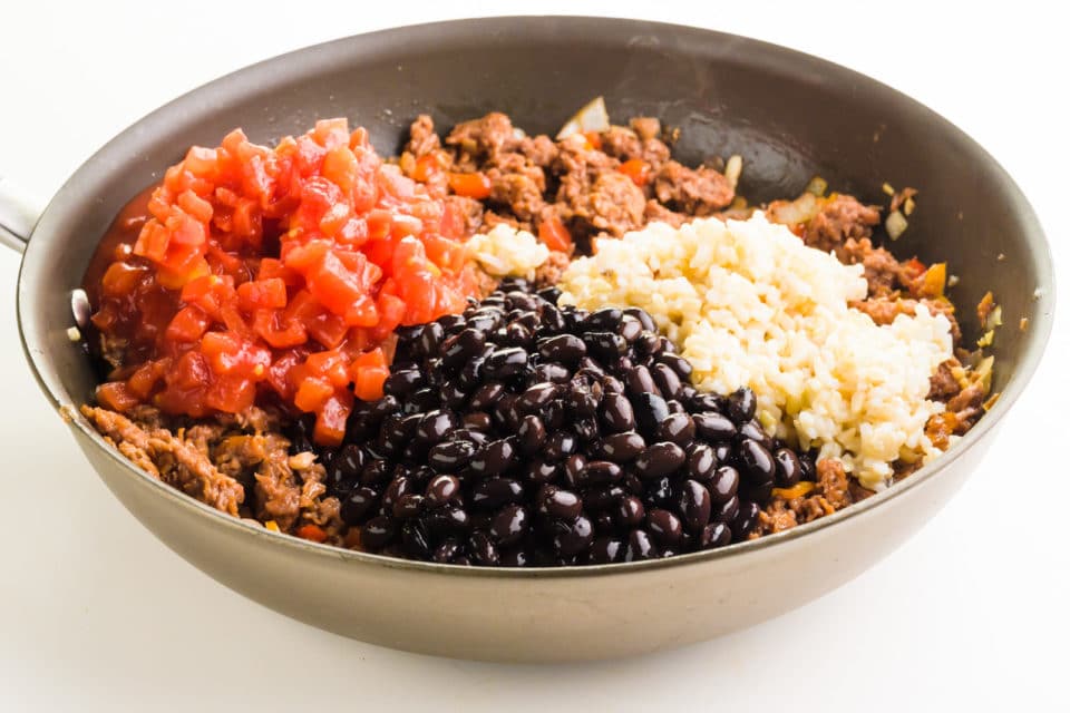 A skillet holds plant-based meat filling, topped with brown rice, black beans, and diced tomatoes.