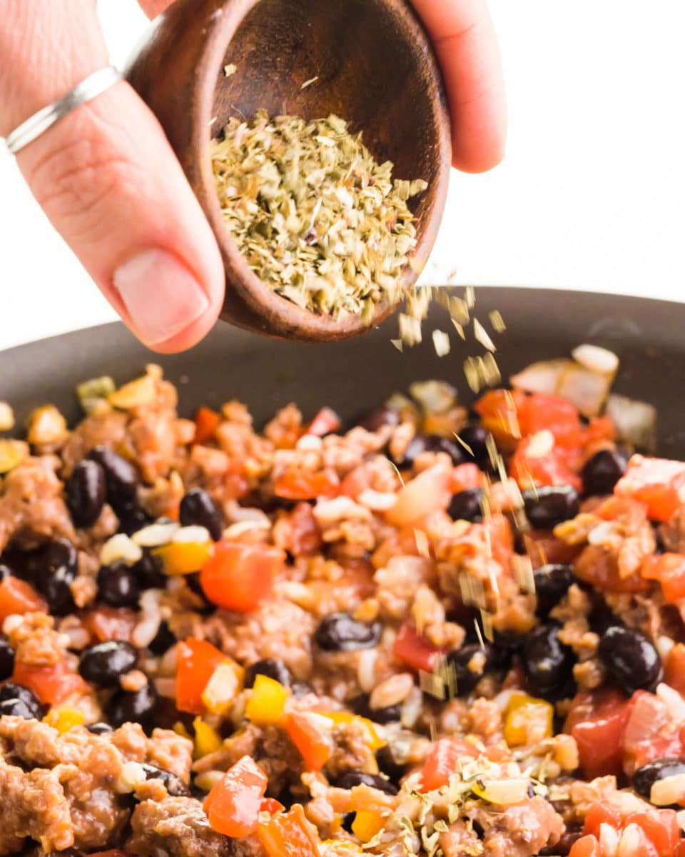 A hand holds a bowl of Italian seasonings and is drizzling over a plant-based meat filling mixture.