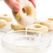 A hand holds a donut, dipping it in vanilla glaze. There are donuts on a wire rack in the background.