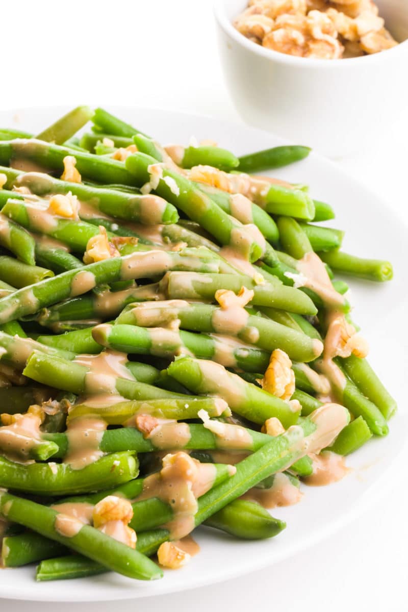 Balsamic green beans have chopped walnuts and glaze over the top. There's a bowl with walnuts in the background.