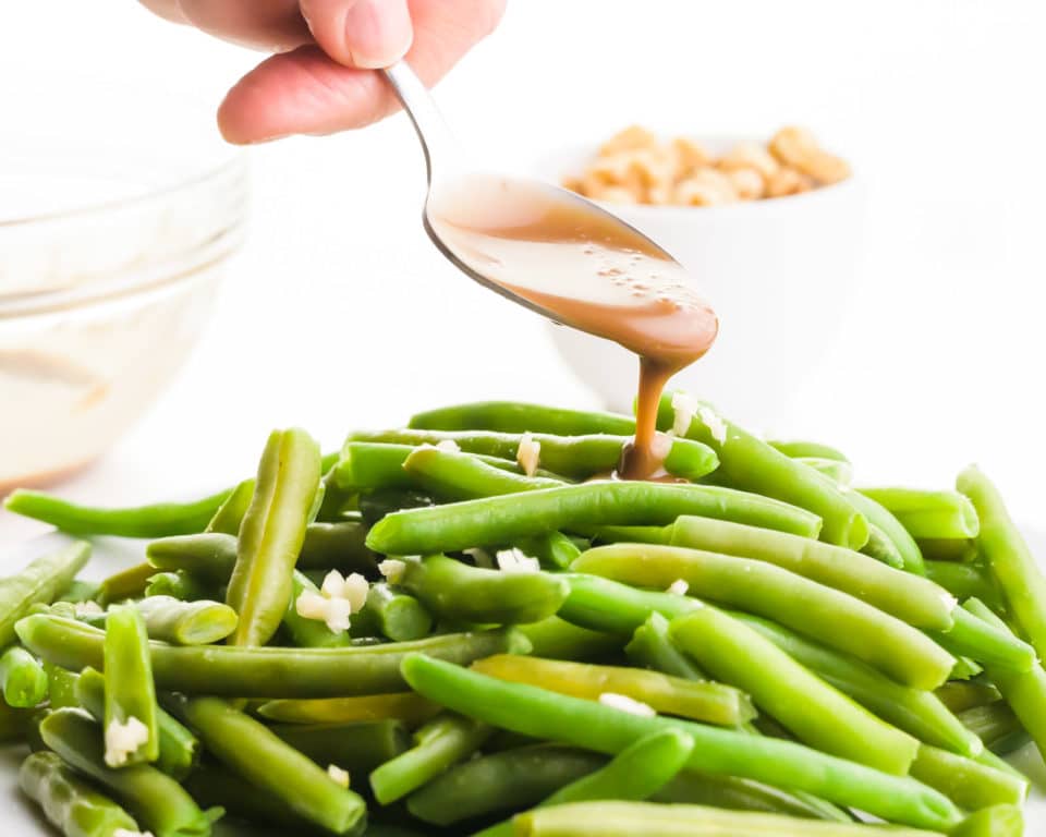 A hand holds a spoon drizzling balsamic glaze over steamed geen beans.