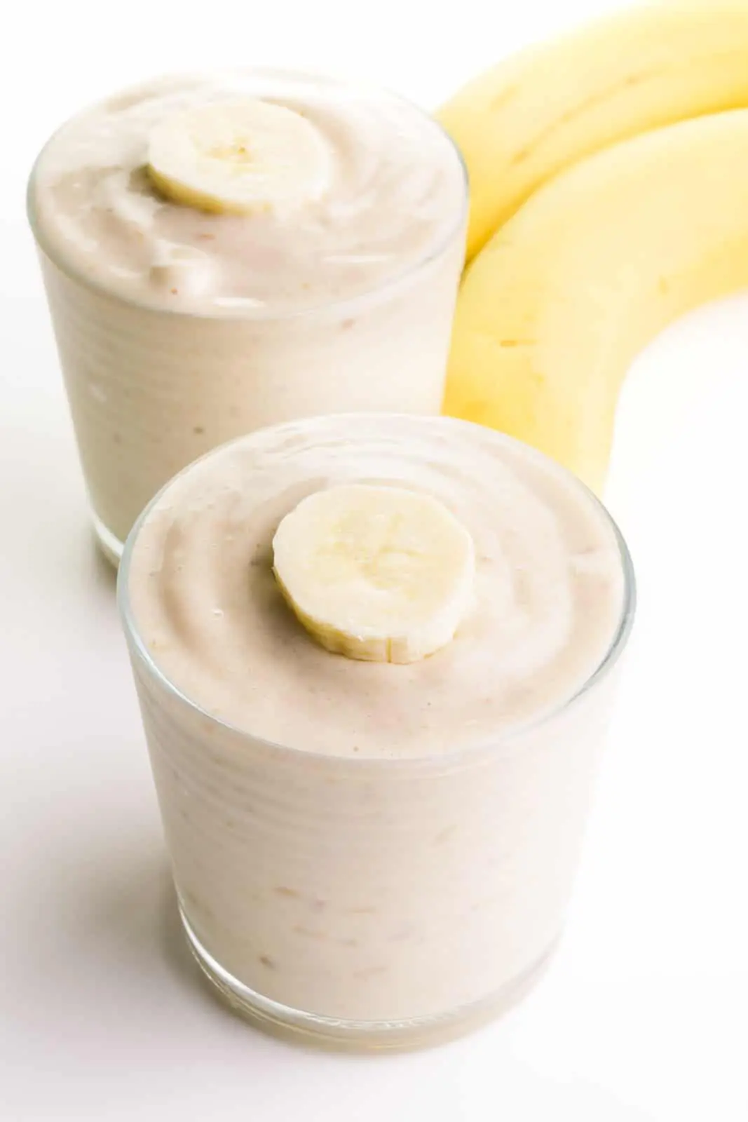 A banana milkshake is in a glass with a slice of banana on top. There's a second glass and some bananas in the background.