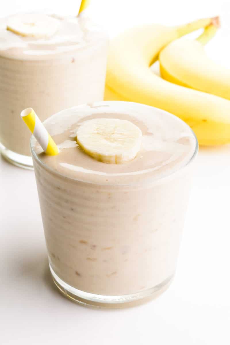 A glass holds some healthy banana shake with a yellow straw on the side. There's another glass in the background and two bananas.