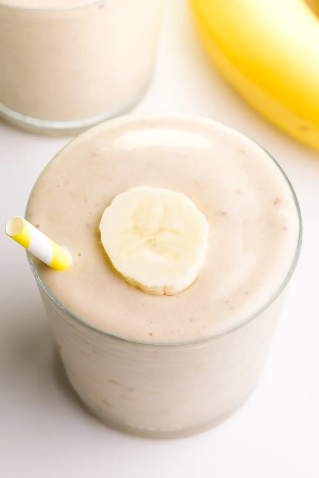 Looking down on a glass with banana smoothy. There's a banana slice on top and a yellow striped straw. There's another glass and a banana in the background.