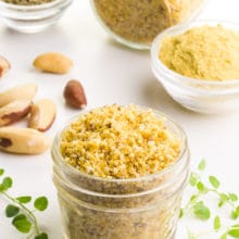 A small glass container of Brazil nut Parmesan has herbs around it. There are Brazil nuts and bowls of ingredients in the background.