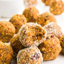 A group of carrot cake energy bites sits around a bowl of walnuts. One of the treats has a bite taken out of it.