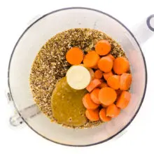 A food processor bowl has ground ingredients in it along with nut butter and carrot slices.