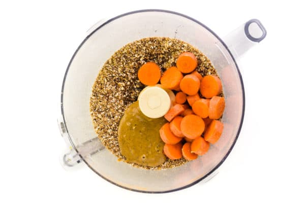A food processor bowl has ground ingredients in it along with nut butter and carrot slices.