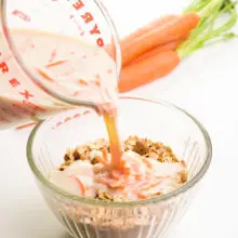 A hand holds a measuring cup pouring wet ingredients into a bowl with oats and spices. There are raw carrots in the background.